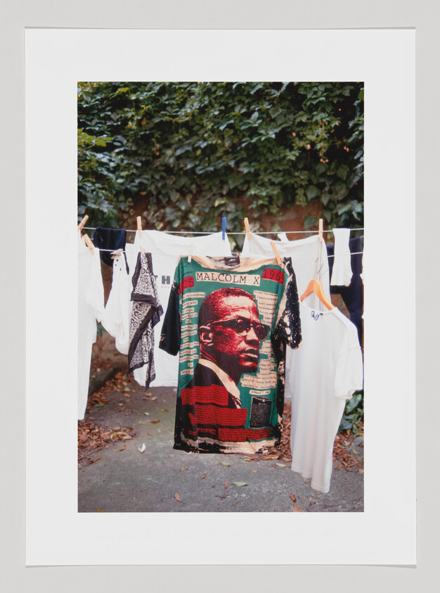 A colorful T-shirt with a graphic of Malcolm X hanging on a clothesline among other laundry items, with a backdrop of lush green foliage.
