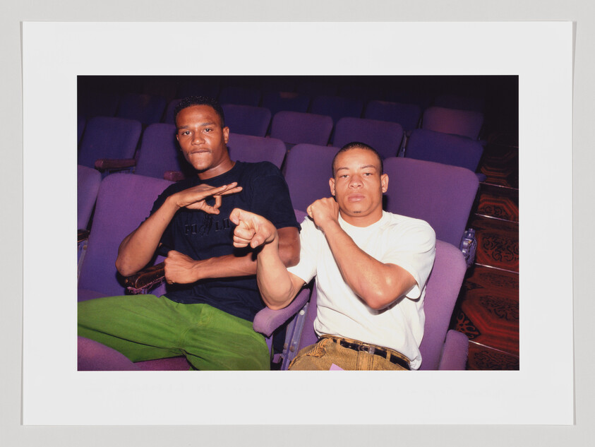 Two men are sitting side by side in a movie theater with purple seats, both making hand gestures towards the camera. The man on the left is wearing a black t-shirt and bright green pants, while the man on the right is in a white t-shirt and patterned brown pants. They appear to be posing for the photograph in a casual and friendly manner.