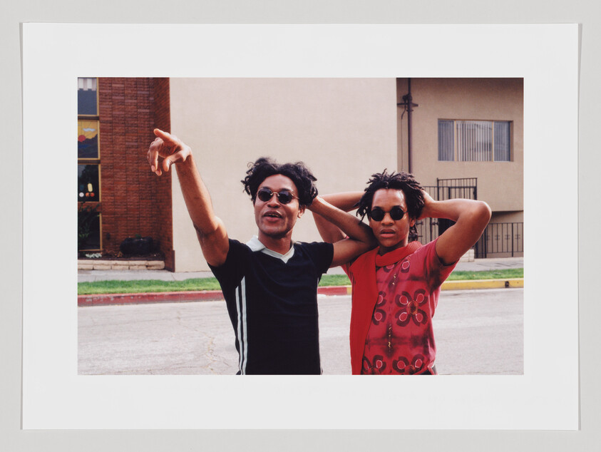 Two individuals stand in the middle of a street, with one person pointing to something out of frame and the other with hands behind their head. Both are wearing sunglasses and casual clothing, with a building and a clear sky in the background.