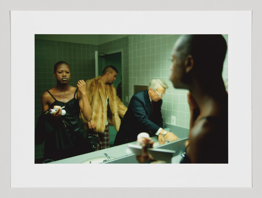A Black person doing their make-up in a public bathroom; the mirror reflects two additional people