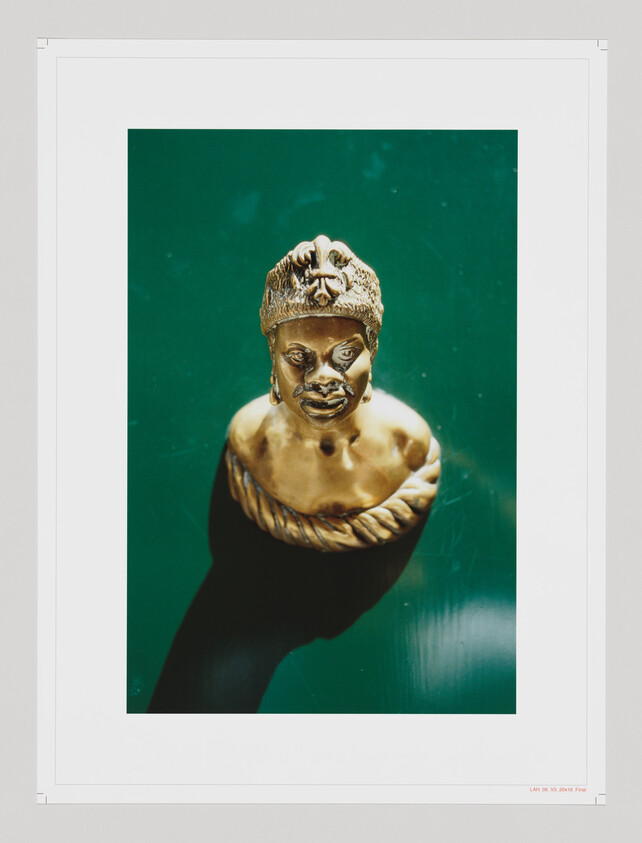 A golden bust of an African figure with detailed facial features and traditional headgear, centered against a teal background with a white border framing the image.