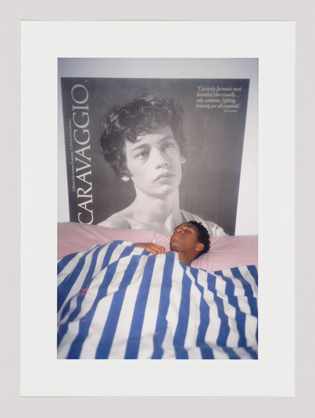 A large magazine cover poster on a wall, in front, a Black man sleeping in a bed with striped covers and pink pillows.