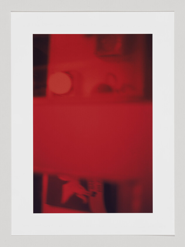 A monochromatic red-toned image with blurred shapes and indistinct forms, giving an abstract or out-of-focus appearance, mounted on a white background.