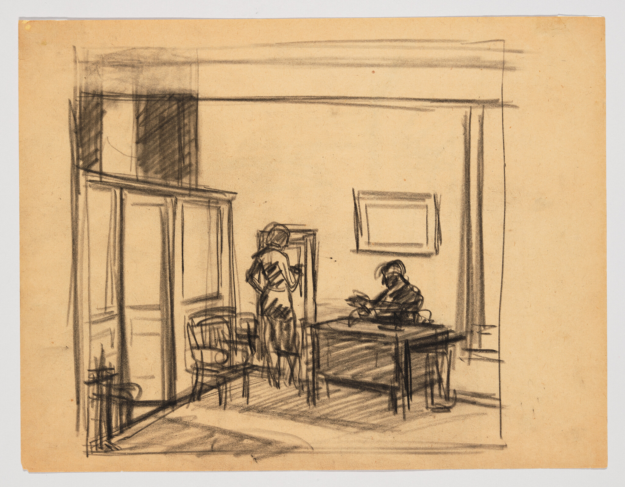 Two people in a room, one standing near a chair and the other sitting at a desk writing.