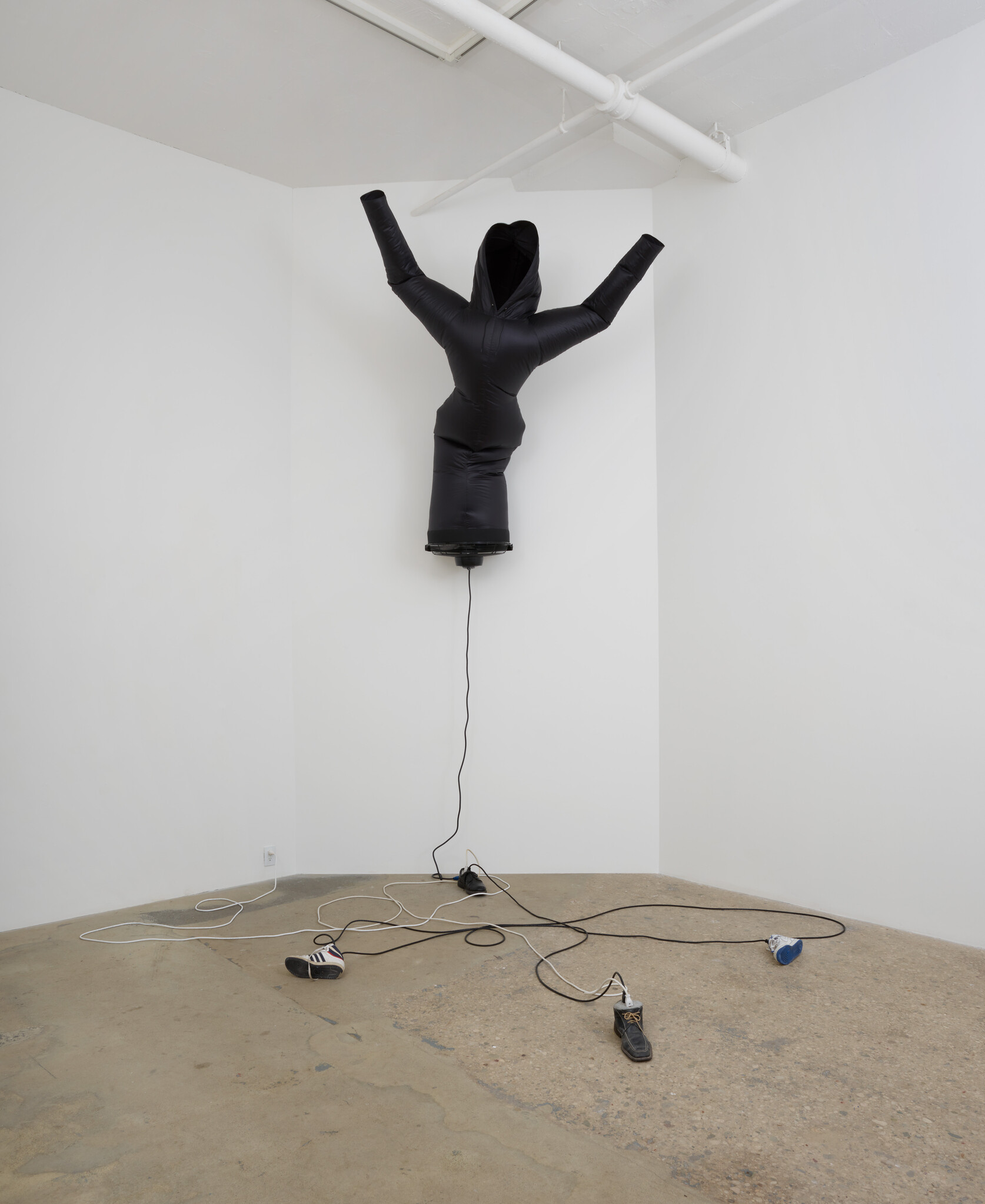 A black inflatable tube man with a humanoid shape is suspended from the ceiling in a white gallery space, with its arms raised and head pointing down. The figure is connected to a black box on the ceiling by a cable, and there are four blowers on the ground with cables running to the suspended figure.