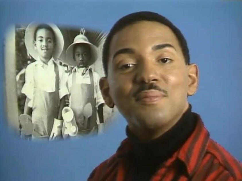 A smiling man in a striped shirt poses beside a faded photograph of two children in costumes.