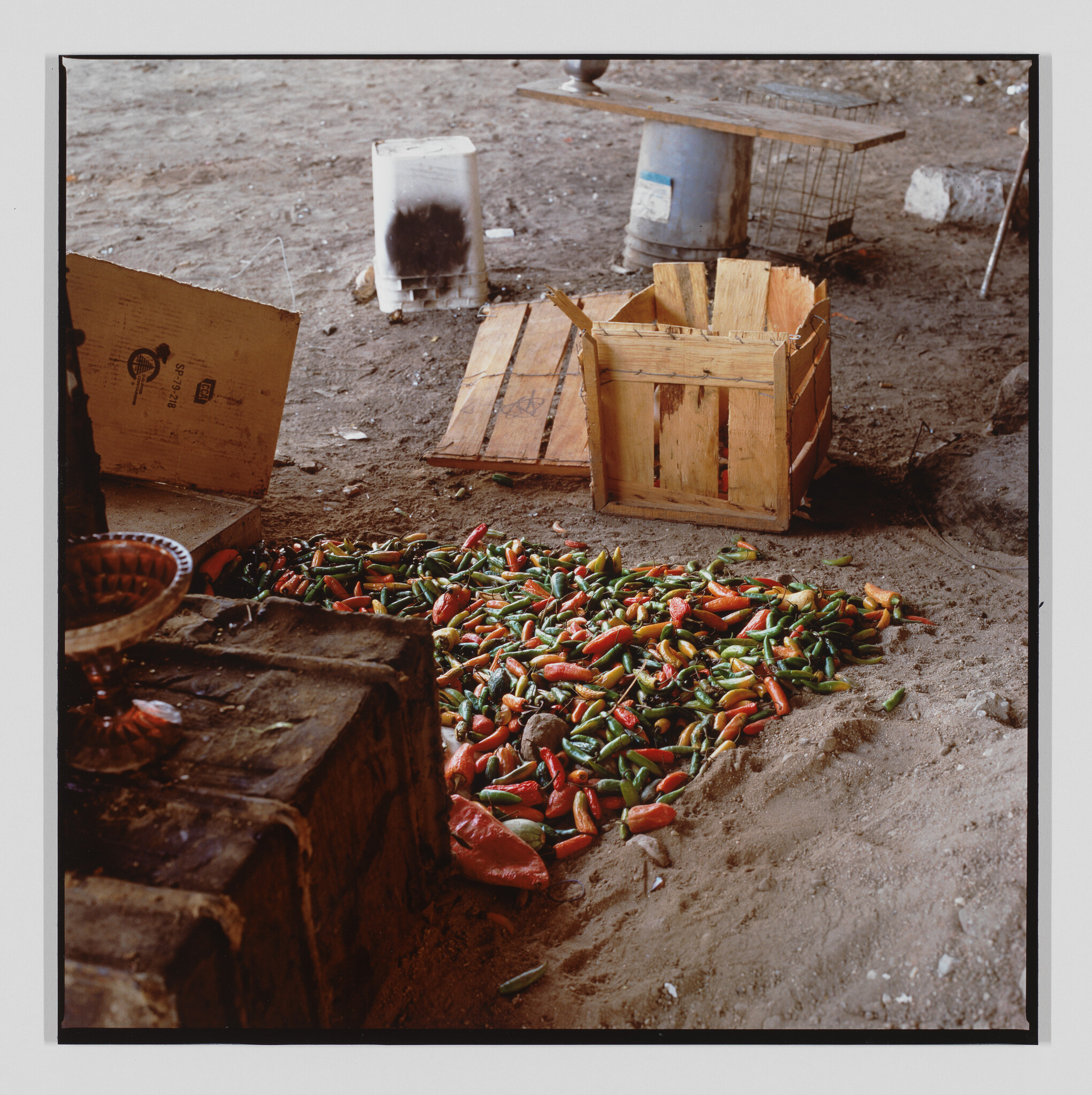 A large pile of red and green chili peppers scattered on a dirt floor next to wooden crates.