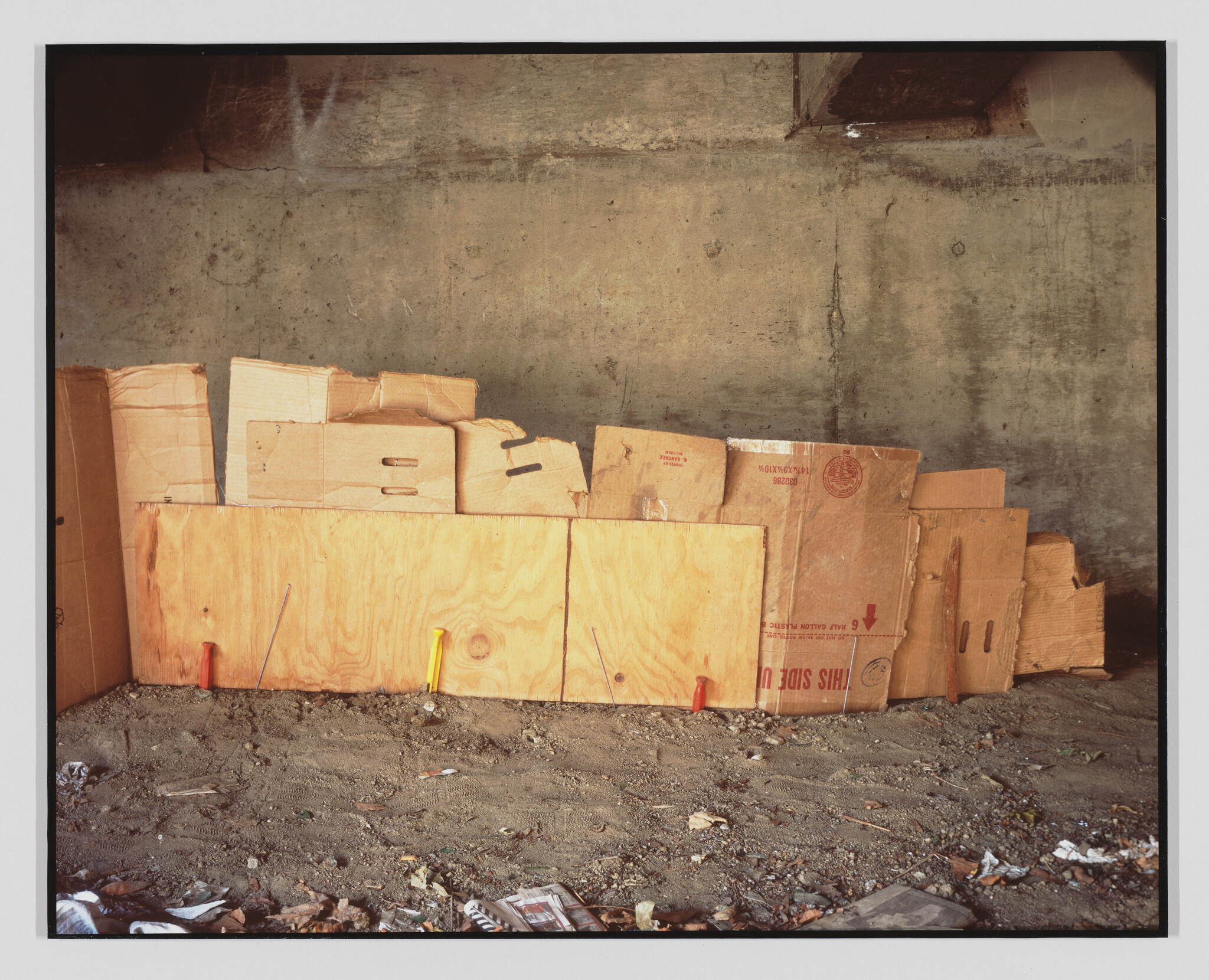 A makeshift wall of cardboard and plywood leans against a concrete underpass over a dirt floor.