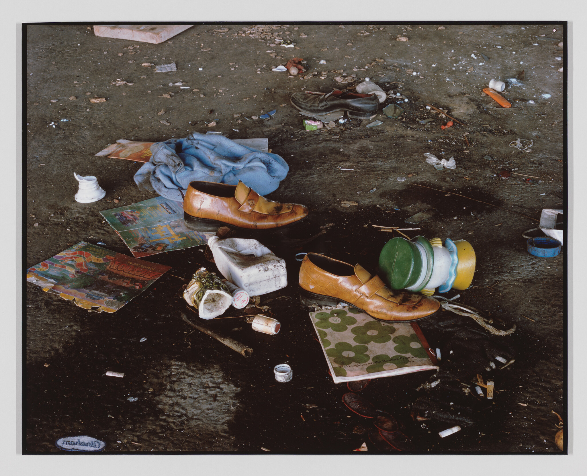 A pair of worn brown dress shoes lies among scattered magazines, containers, and trash on a dirty floor.