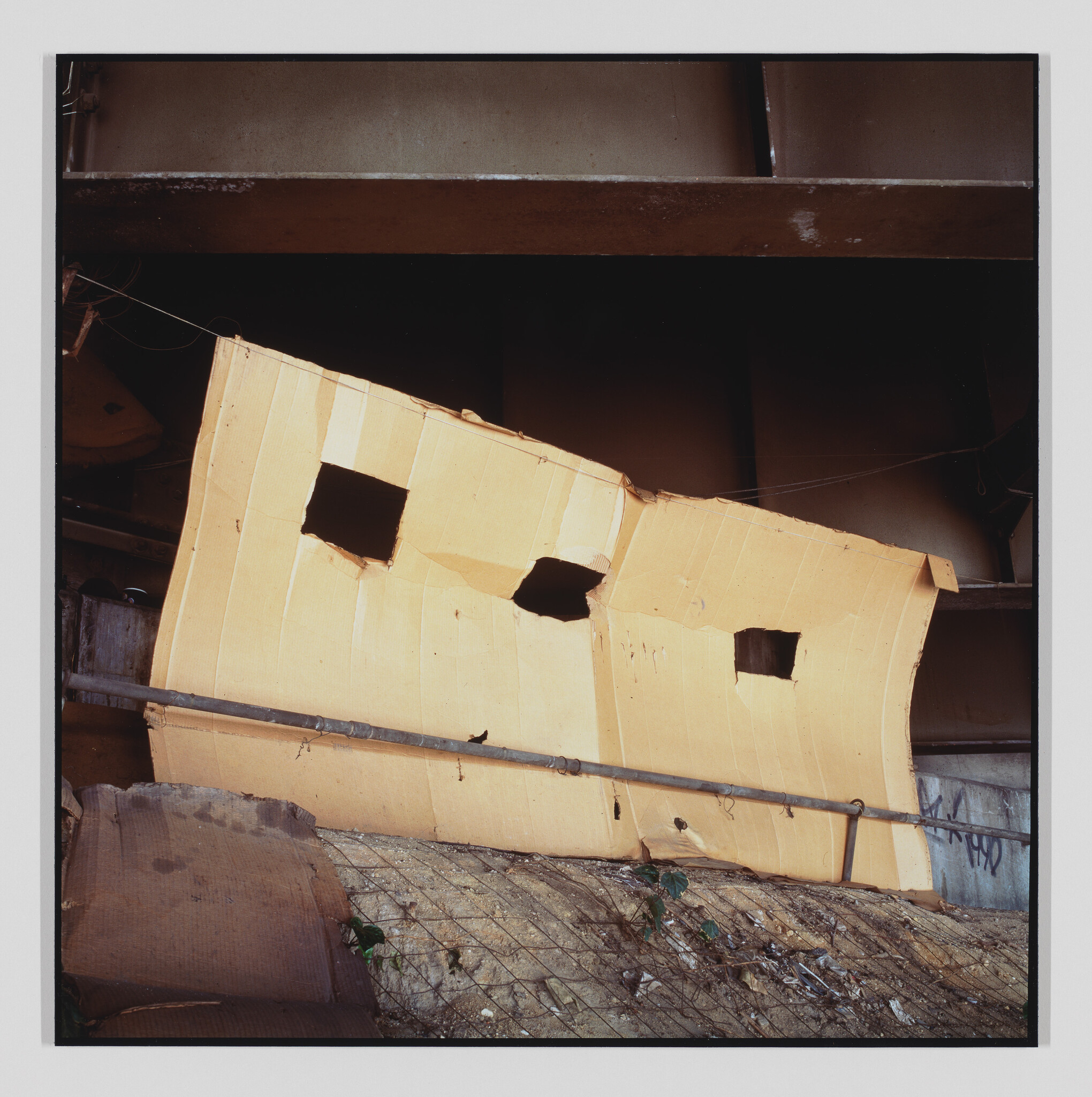 Large torn cardboard with three square holes propped against a metal railing under a bridge.