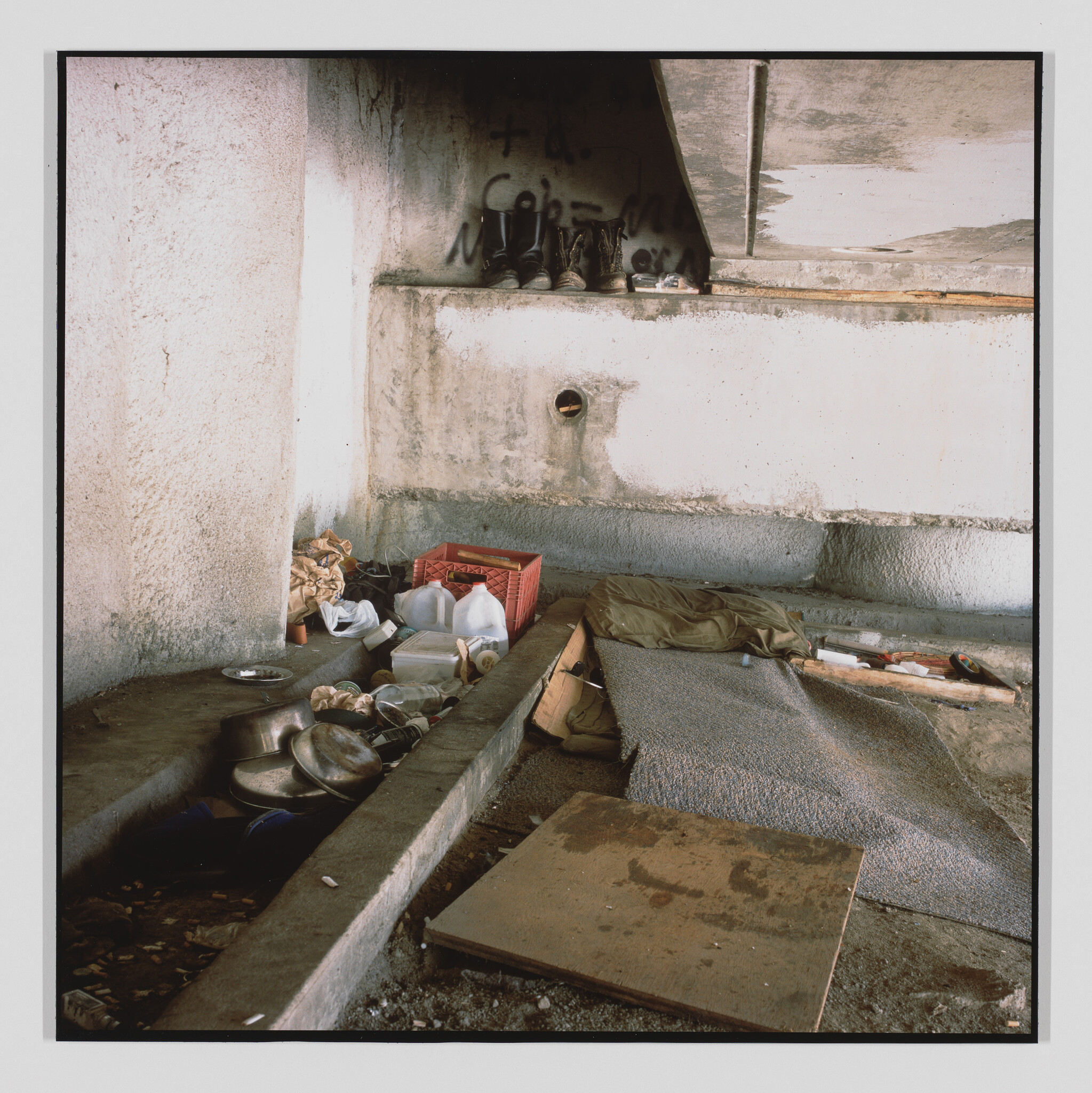 Makeshift sleeping area under a concrete overpass with scattered pots, jugs, boots, and a worn blanket.