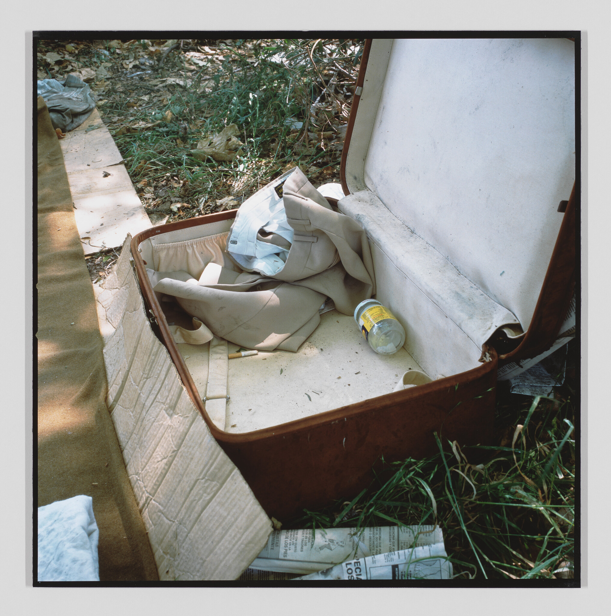 An open suitcase lies in grass containing folded beige clothes, a plastic jar, and a newspaper.