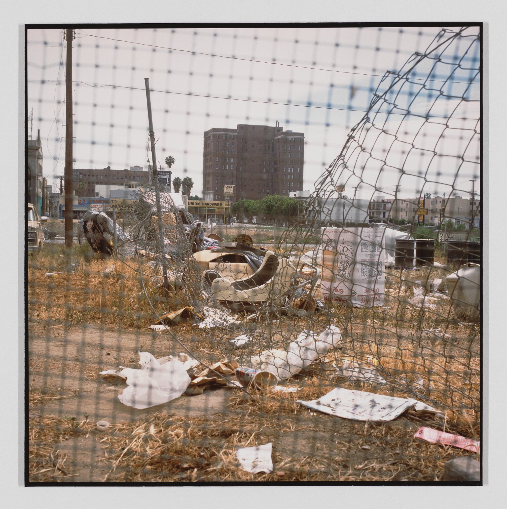 A torn chain-link fence surrounds a trash-filled vacant lot with discarded car parts and debris.