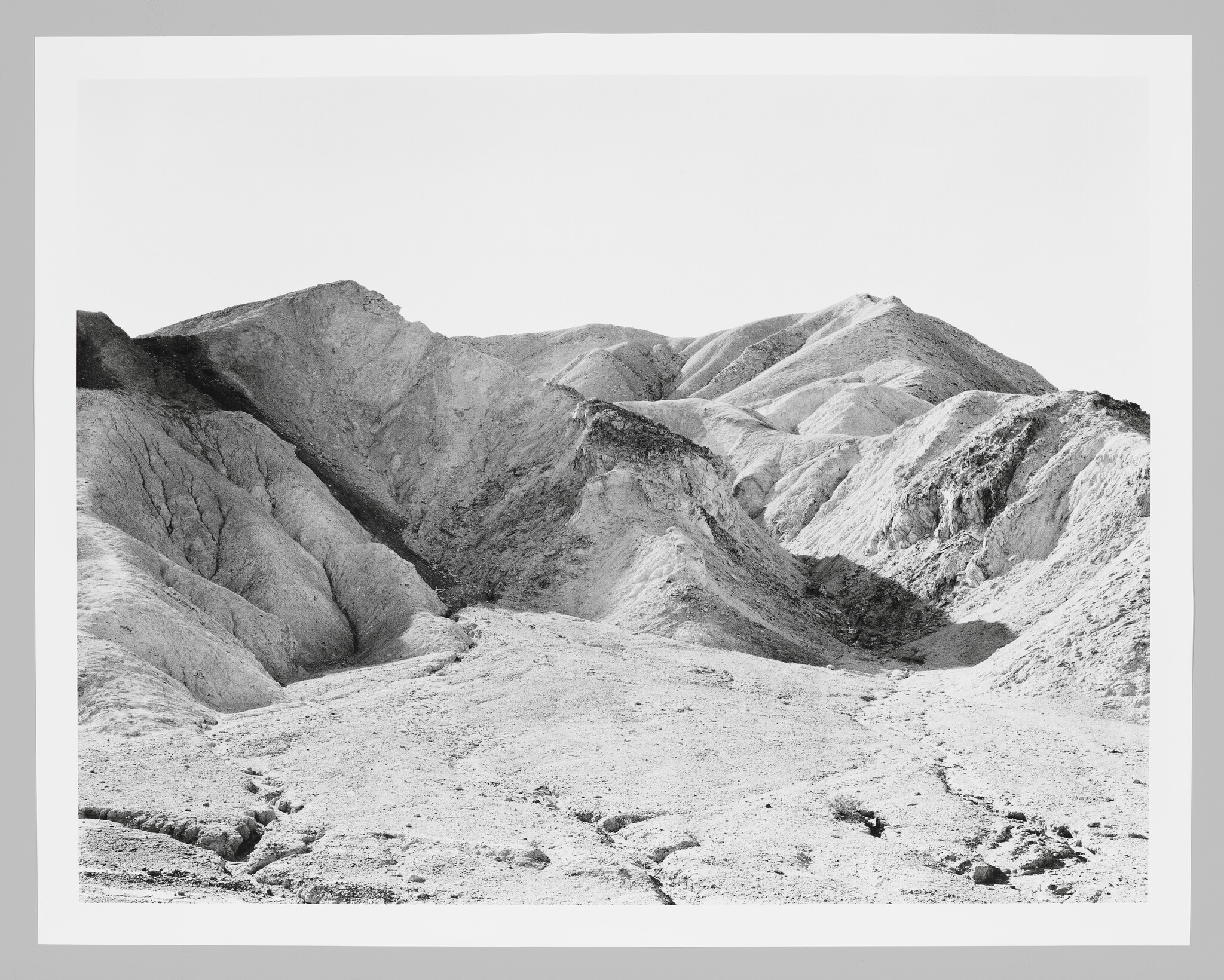 Barren eroded hills rise from cracked dry ground under a clear sky.