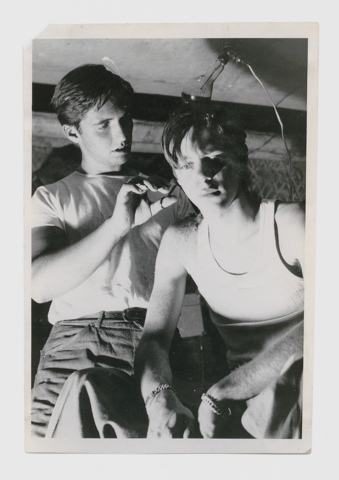 A young man cuts another young man's hair while both sit under a hanging light.