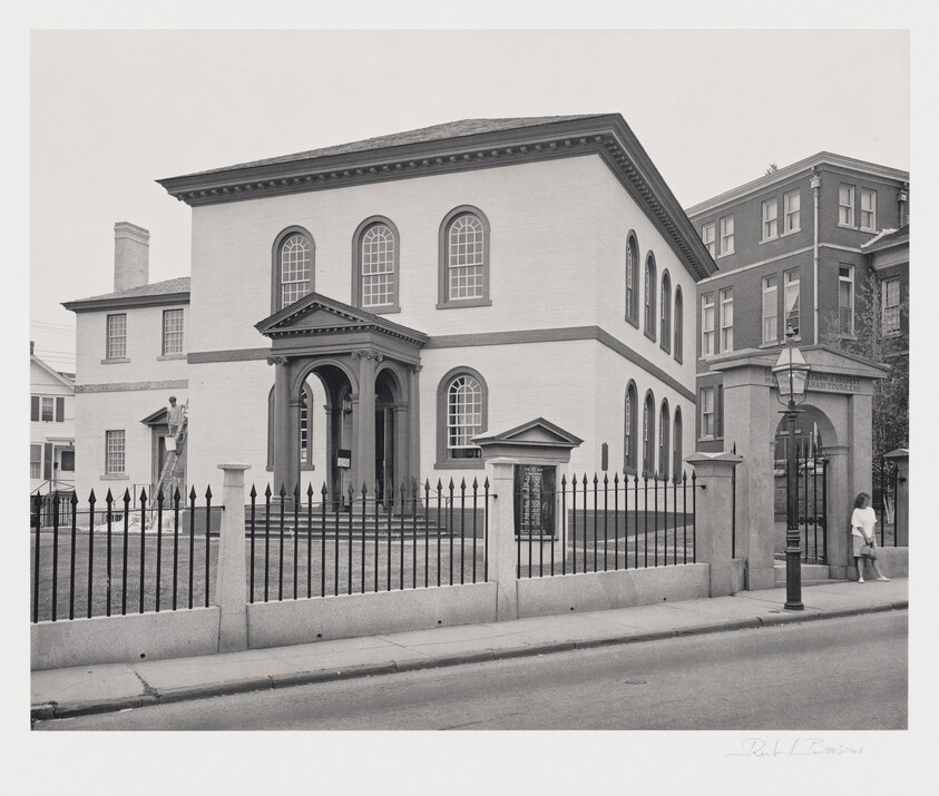 A black and white photograph of a historic two-story building with a classical facade, featuring large arched windows on the second floor and a portico with columns at the entrance. A wrought iron fence surrounds the property, and there is a street lamp post near the sidewalk. In the background, other older buildings are visible, and a person is seen walking by the fence on the right side of the image. The photo is signed by the photographer at the bottom right corner.