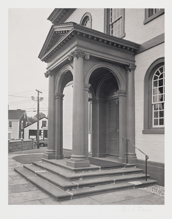 A black and white photograph of the entrance to a classical building with a portico supported by columns, a set of stone steps leading up to a wooden door, and a metal handrail on the right. In the background, residential houses and utility poles are visible. The image is signed at the bottom right corner.