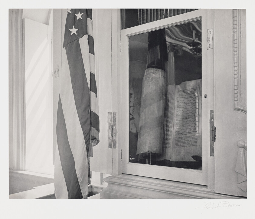 A black and white photograph showing an American flag hanging vertically next to a window with a reflection of the flag. Through the window, a framed document is visible, possibly of historical significance, mounted on a wall inside the room. The image has a signature in the bottom right corner.