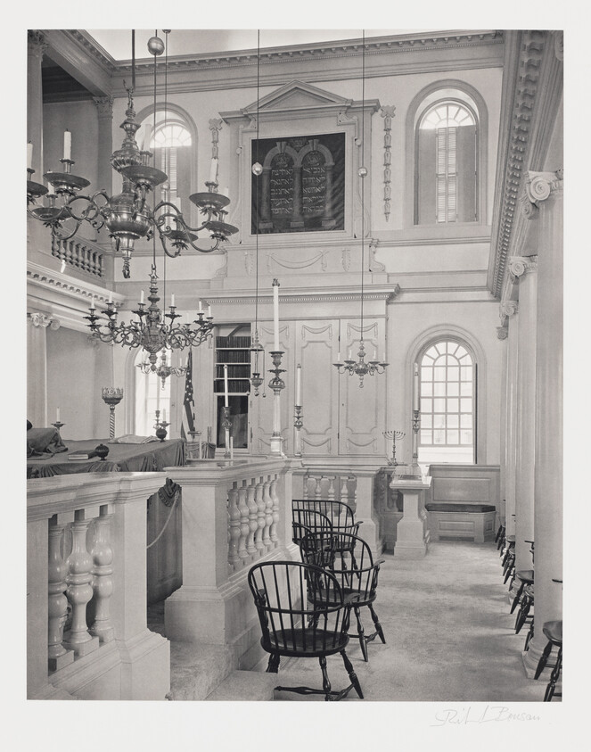 Black and white photograph of an elegant interior, likely a synagogue, featuring a balcony with a balustrade, several ornate chandeliers, and a wall with Hebrew inscriptions, possibly the Ten Commandments. Wooden chairs are arranged in the foreground, and natural light streams in through arched windows.