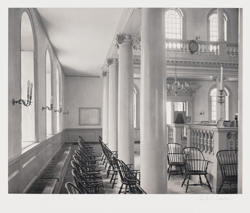 Black and white photograph of an elegant interior with classical architectural details, featuring rows of empty chairs facing a pulpit, flanked by tall columns and arched windows with wall-mounted candle sconces. A balcony with a balustrade is visible in the background, and a chandelier hangs from the ceiling. The image conveys a serene and historical atmosphere.