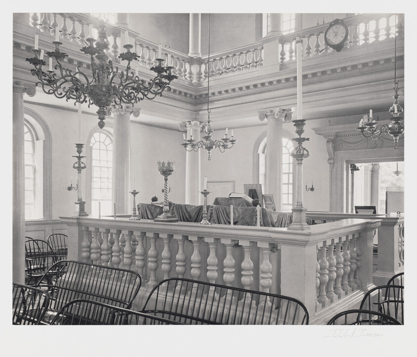 A black and white photograph of an elegant, historic courtroom interior with rows of empty chairs facing a judge's bench. The bench is elevated and adorned with a draped cloth. Ornate chandeliers hang from the ceiling, and classical columns and a balcony with a balustrade are visible in the background. A clock is mounted on the wall near the balcony.