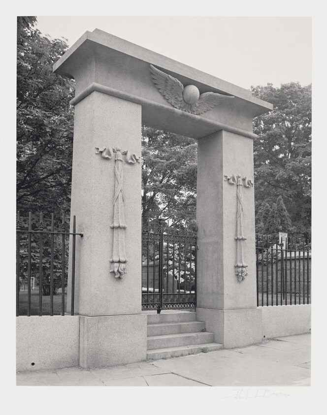 A black and white photograph of a stone memorial archway with decorative elements including a winged globe at the top and floral garlands on the sides. The archway is flanked by a metal fence and is situated in front of a backdrop of trees. There are steps leading up to the gated entrance under the arch. The image is signed at the bottom right corner.
