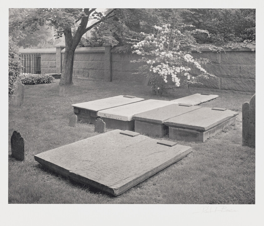 A black and white photograph of an old cemetery with several flat, weathered gravestones and headstones on a grassy ground. A flowering bush with white blooms is visible against a stone wall in the background, and there's a wrought iron gate partially seen to the left. The image has a signature on the bottom right that appears to be "Robt Benson."