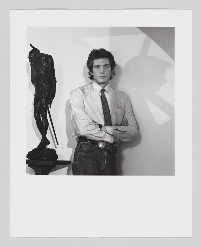 A man in a shirt and tie stands with arms crossed beside a tall statue casting a shadow.