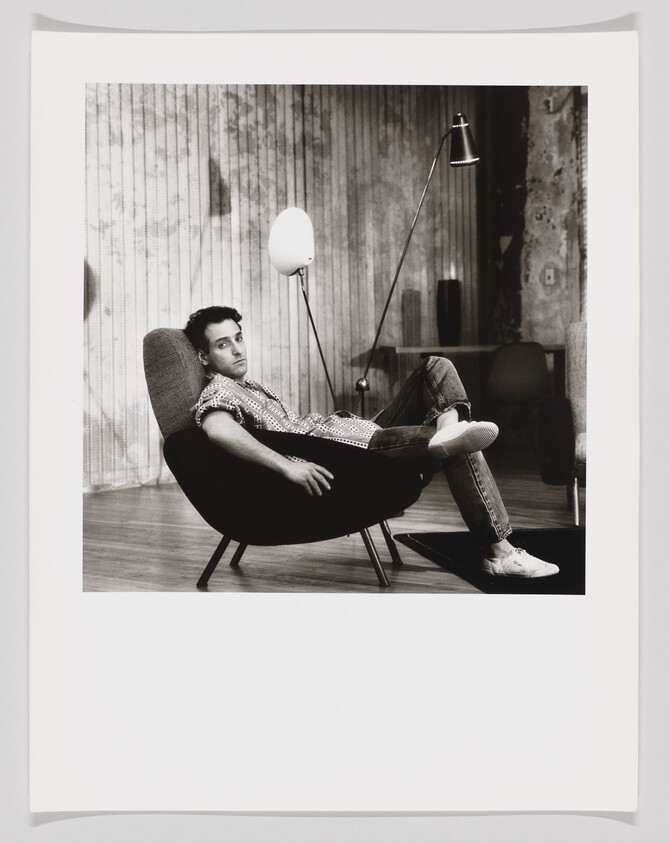 A young man lounges in a modern chair with one leg crossed, looking toward the camera.