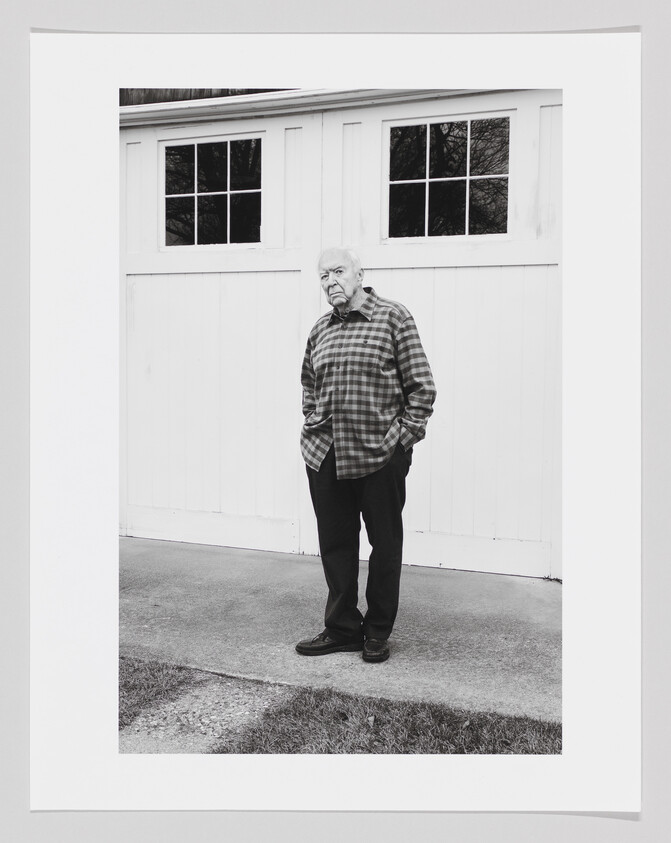 A black and white photo of Jasper Johns in a flannel standing with hands in his pockets.