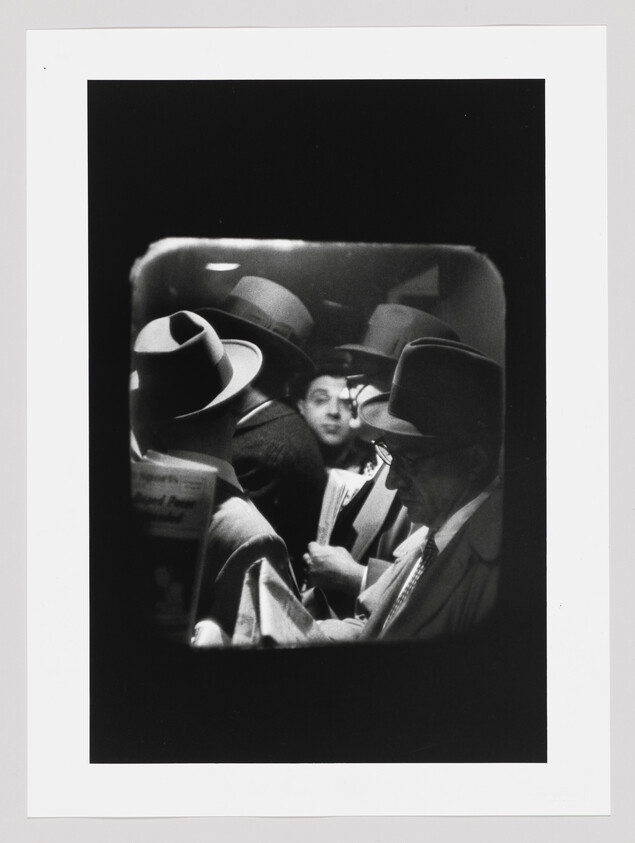 Several men wearing hats stand close together in a crowded subway car, one reading a newspaper.