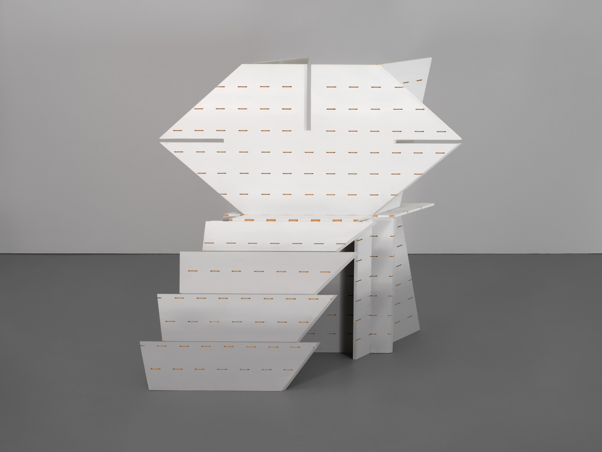 White geometric sculpture made of interlocking slotted panels stands on a gray floor in a gallery.