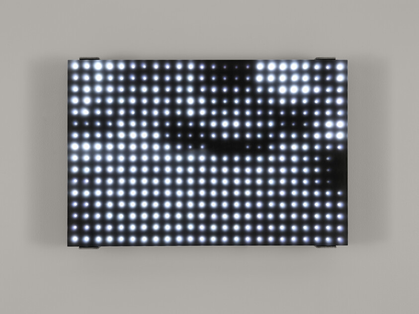 A large digital display composed of numerous bright white LED lights arranged in a grid pattern, with a darkened area in the center creating a shadow effect, mounted on a light-colored wall.