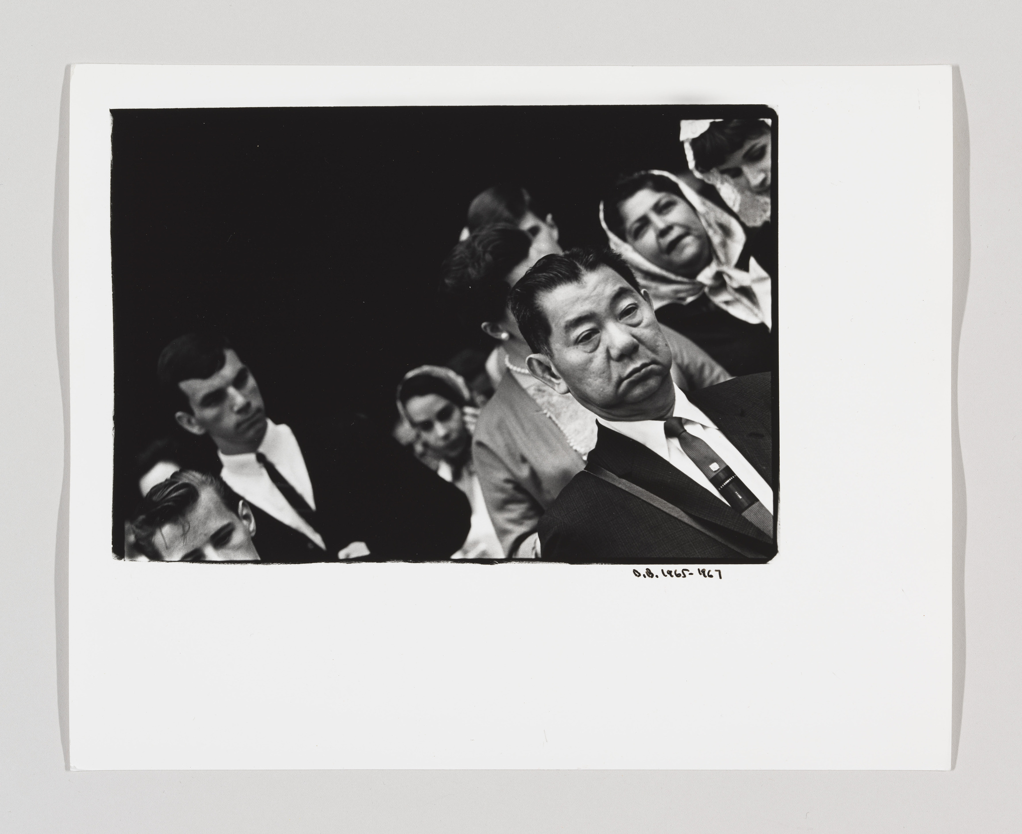 Black and white photograph of a group of people at an event, with a man in a suit in the foreground looking to the side with a serious expression. The image is framed with a white border and has a handwritten inscription at the bottom right corner.