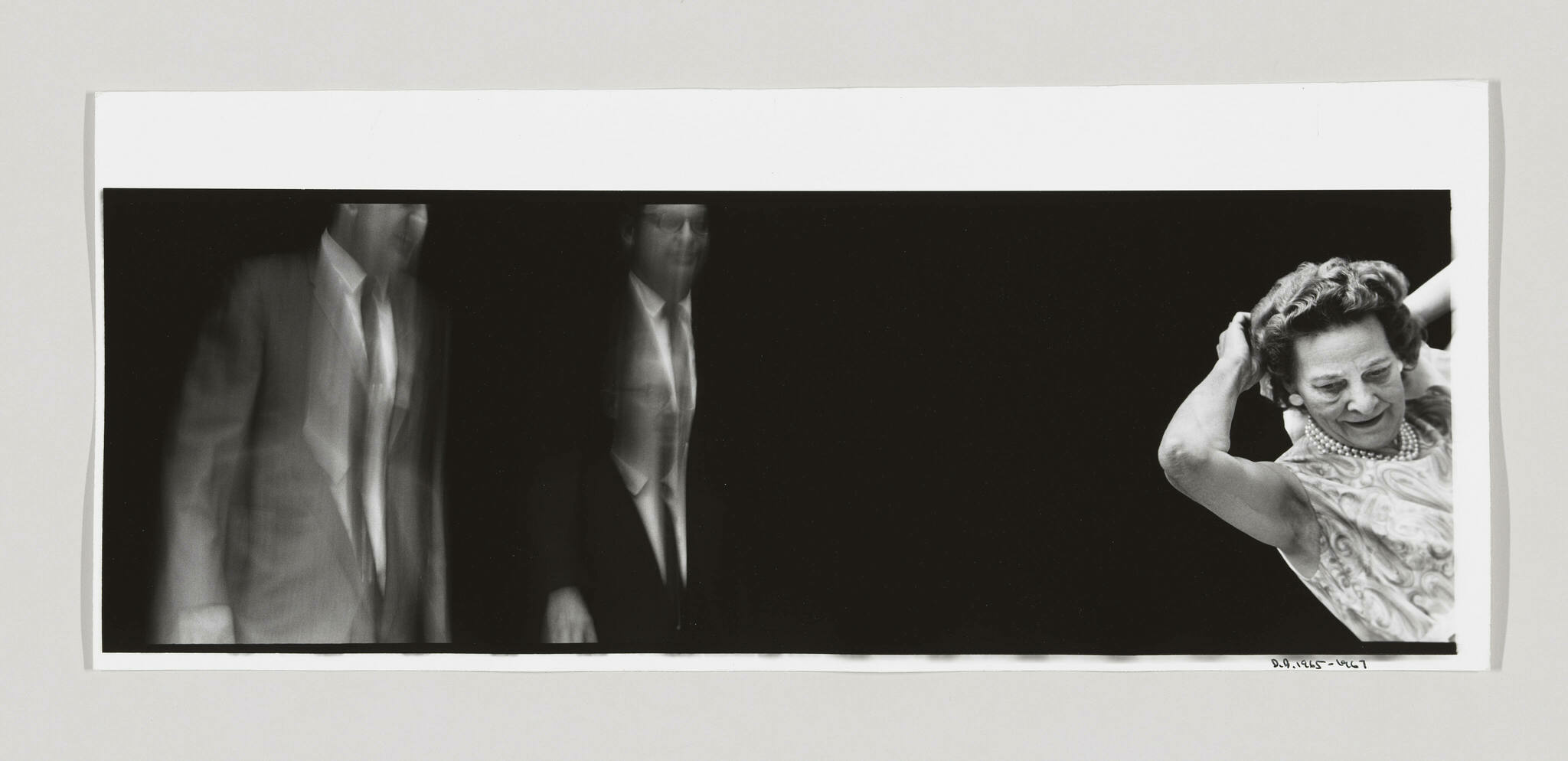 A black and white panoramic photograph featuring blurred images of two men in suits on the left and a clear image of an older woman adjusting her hair on the right, against a black background. The photo is mounted on white paper with the artist's signature and date on the bottom right.