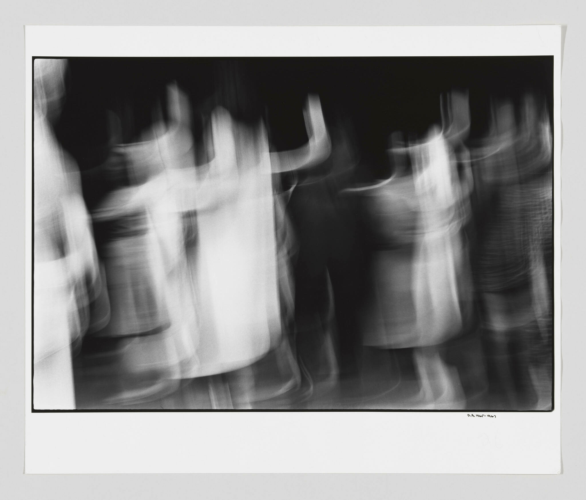 A black and white photograph capturing motion blur of several people, creating abstract, ghost-like figures against a dark background. The image is framed with a white border and has a signature at the bottom right corner.