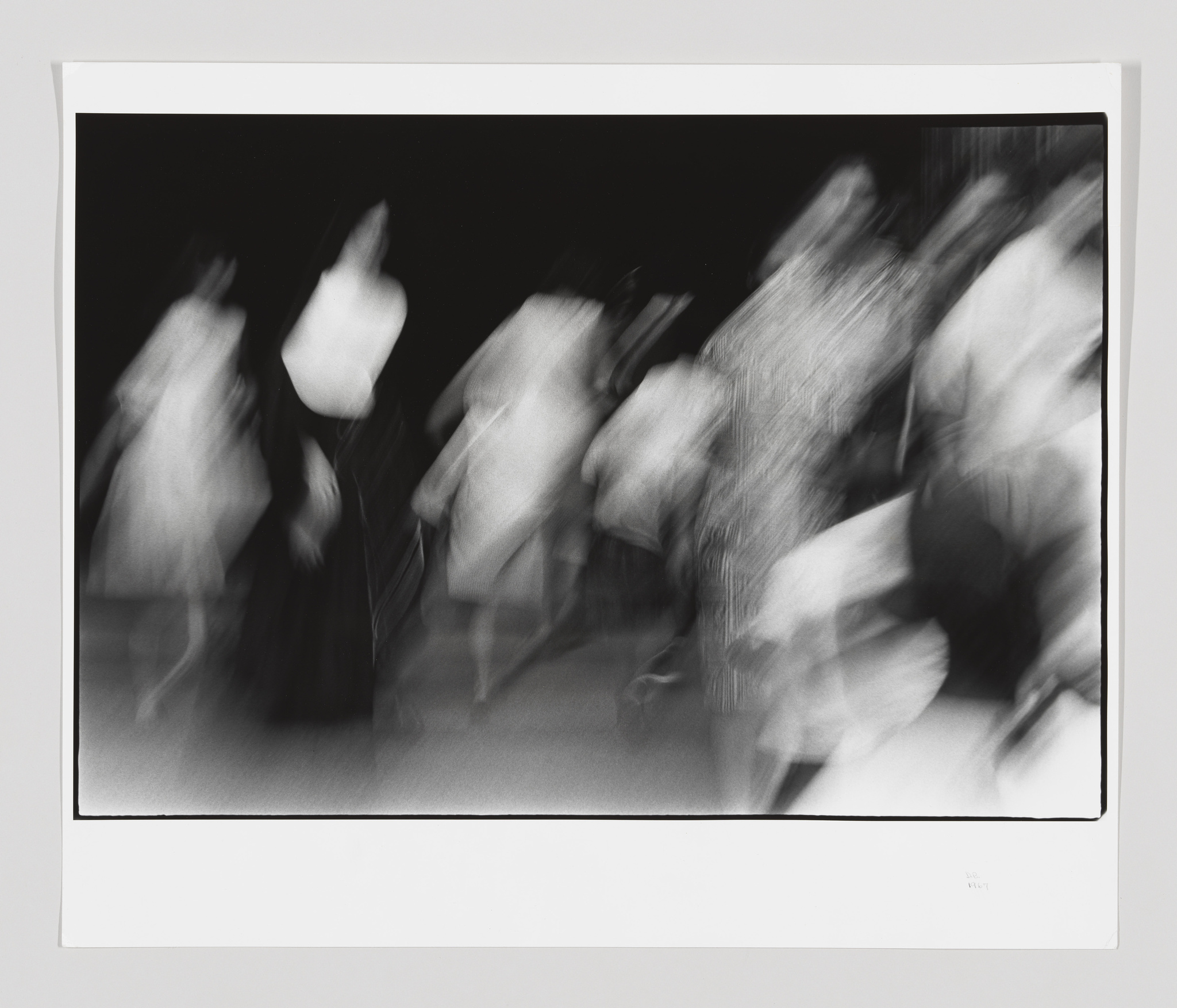 A black and white photograph capturing a blurred motion of people, possibly dancers, in a dynamic and abstract composition, suggesting movement and energy. The image is framed with a white border and appears to be a print with a signature or text in the lower right corner.