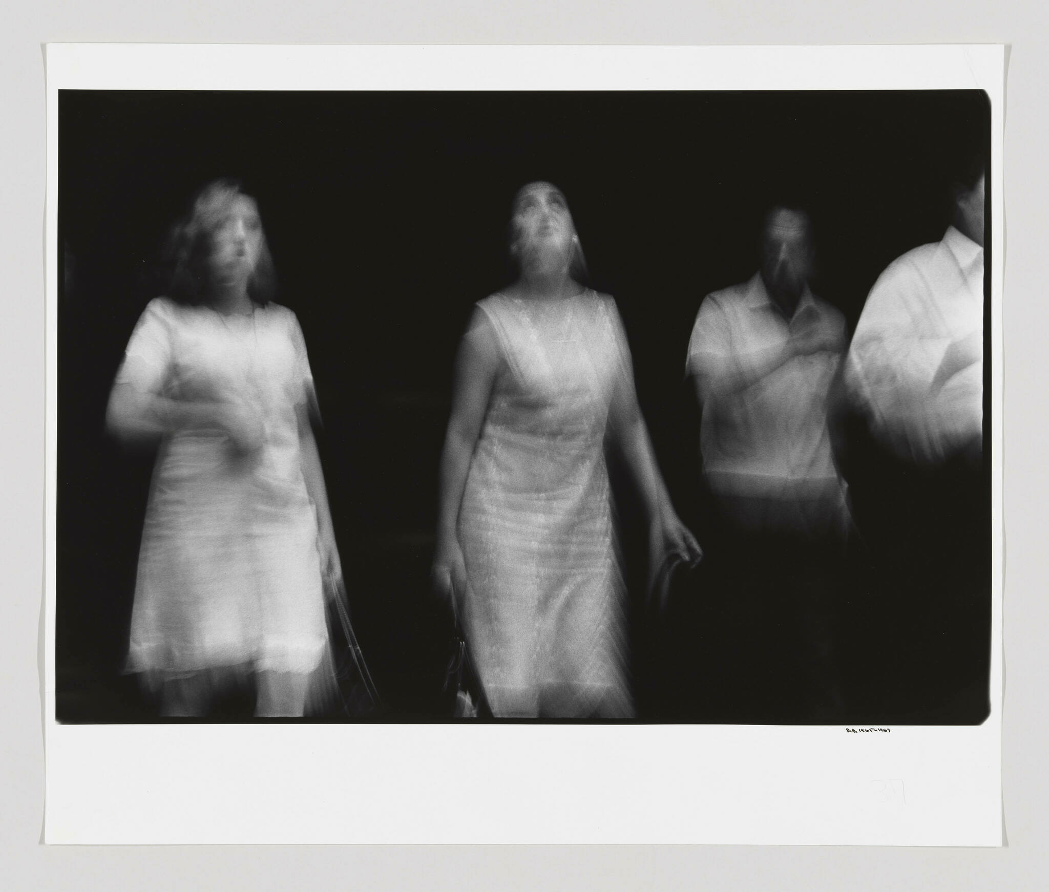 A black and white photograph capturing the blurred motion of two women and two men, giving the impression of multiple exposures or movement during a long exposure. The figures appear ghostlike against a dark background, with the women in dresses and the men in shirts, all in a state of motion or transition. The image is framed with a white border, and there is a signature at the bottom right corner.