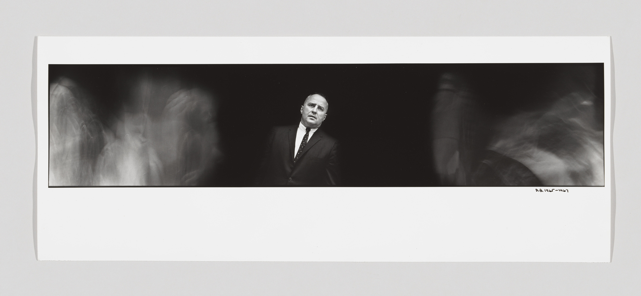 A black and white panoramic photograph featuring a sharply focused man in a suit and tie centered between blurred images of other figures in motion, creating a ghostly effect. The photo is mounted on a white background with a signature and date on the bottom right.