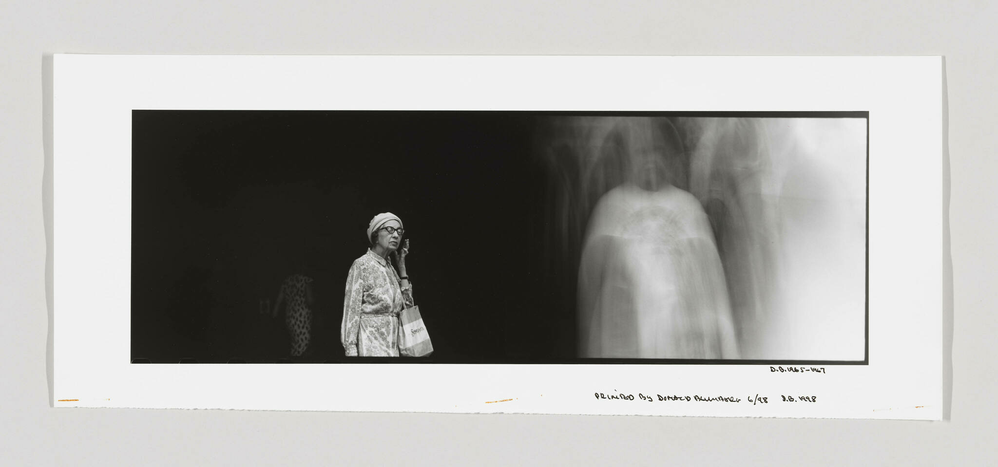 A black and white panoramic photograph featuring a clear image of an elderly woman standing on the left, wearing glasses, a headscarf, and a patterned coat, holding a bag with the word "Safeway" on it. To her right, there is a blurred, ghost-like image of a human figure, resembling an X-ray, against a dark background. The bottom of the image includes handwritten text and numbers, indicating the print number and date.