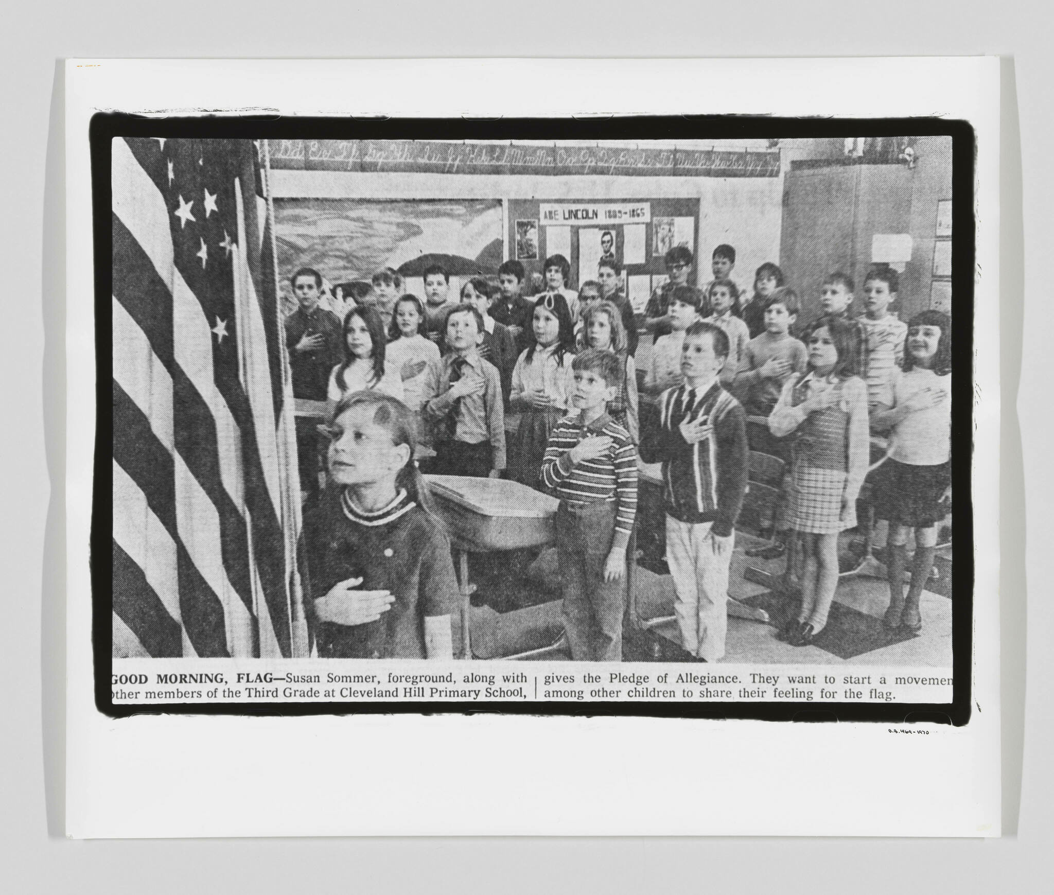 A black and white photo of a group of schoolchildren with their hands over their hearts, facing the American flag to the left as they recite the Pledge of Allegiance in a classroom. One child in the foreground looks towards the flag with a focused expression. The image includes a caption at the bottom that provides context to the scene.