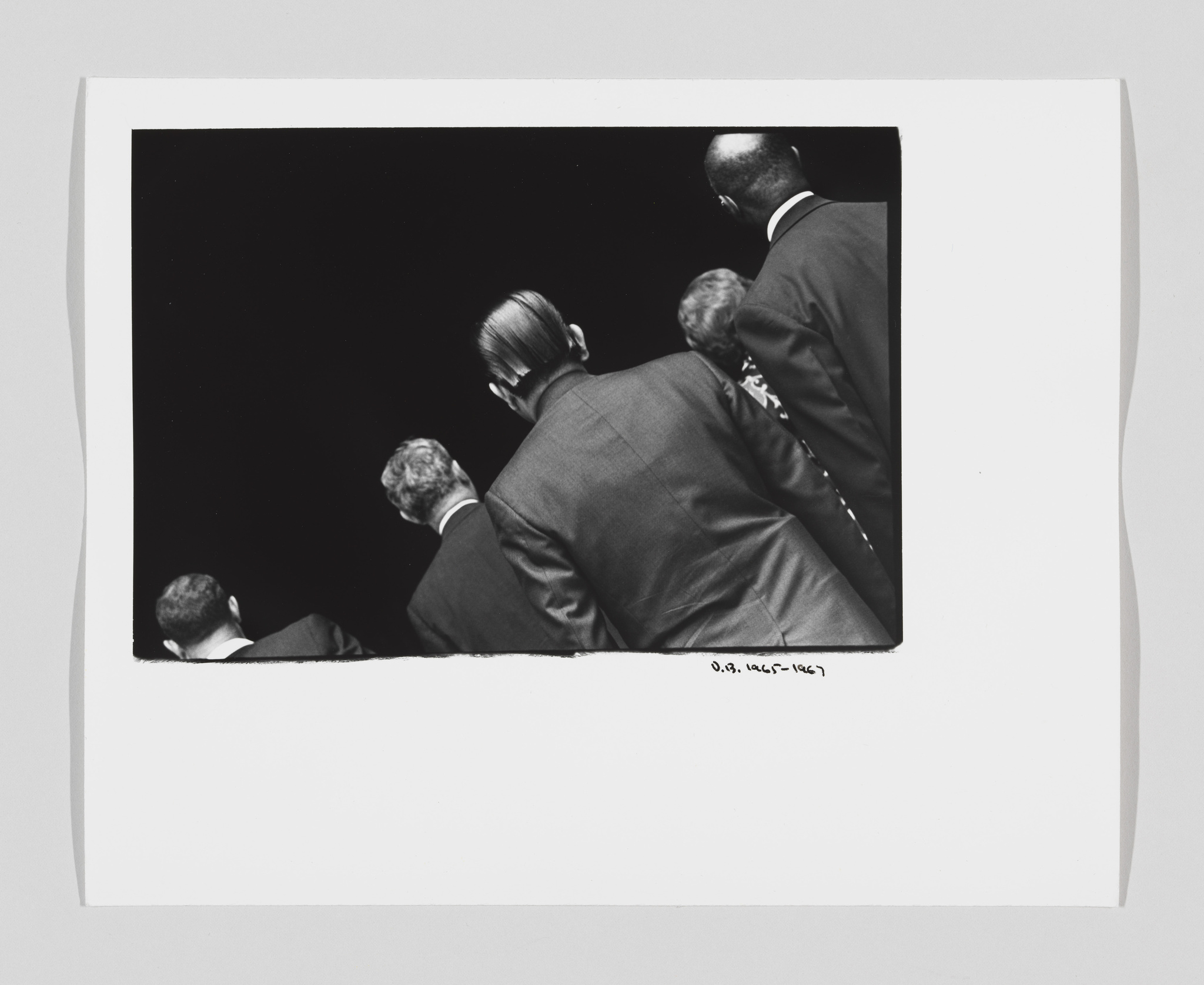 Black and white photograph showing the back view of several men in suits, with a dark background, framed by a white border with handwritten text at the bottom right corner.