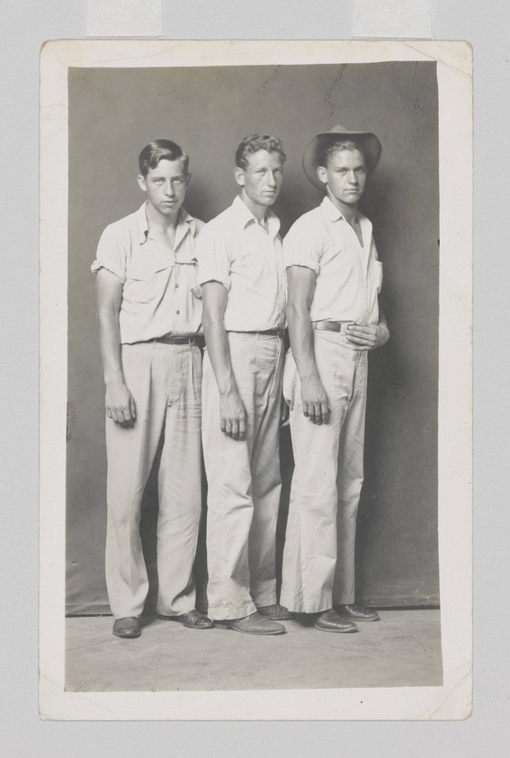 Three young men standing side by side in a vintage black and white photograph. They are dressed in similar light-colored shirts and trousers, with the man on the right wearing a cowboy hat. The photo has a worn white border, indicating age, and shows some signs of damage and creasing.