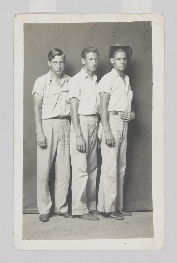 Three young men stand closely in light shirts and trousers, the rightmost wearing a hat.
