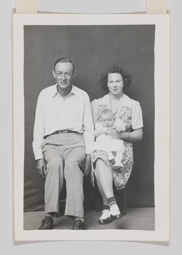 A black and white photograph of a smiling man and woman seated side by side with a baby on the woman's lap. The man is wearing a light-colored shirt and trousers with dark shoes, and the woman is dressed in a patterned dress with white socks and dark Mary Jane shoes. The baby is wearing a light outfit and socks. They are posed against a plain, dark backdrop, and the photo has a white border.