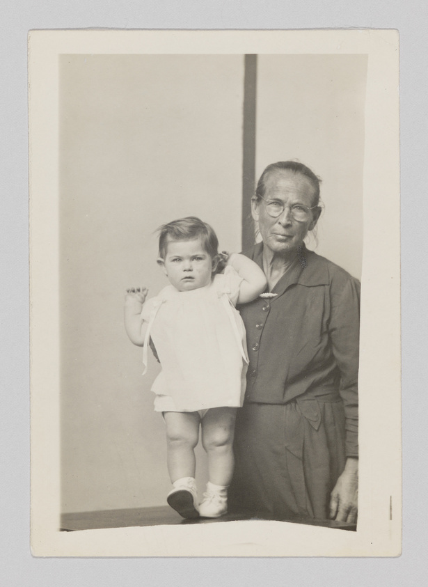 A vintage black and white photograph showing an elderly person standing next to a young child. The elderly individual is wearing glasses and a long-sleeved dress or shirt with buttons, while the child is in a light-colored dress with short sleeves and white shoes. Both are looking directly at the camera, with a neutral background that provides no additional context. The photo has a white border typical of old photographs.