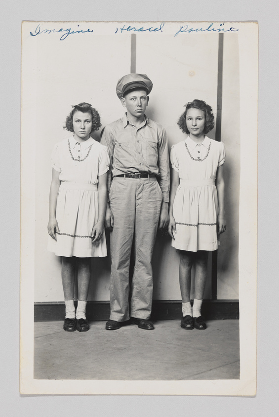 A vintage black and white photograph of two young girls in identical dresses and a young boy in a button-up shirt and trousers, all standing against a plain backdrop. The boy, wearing a cap, stands between the girls, and all three have serious expressions. Handwritten text is visible at the top of the image.