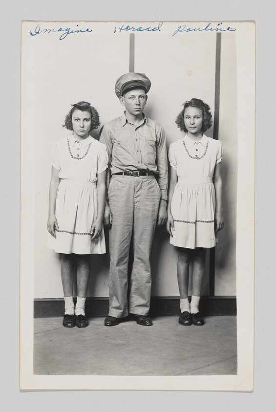 Two girls in matching dresses stand on either side of a boy wearing a cap.