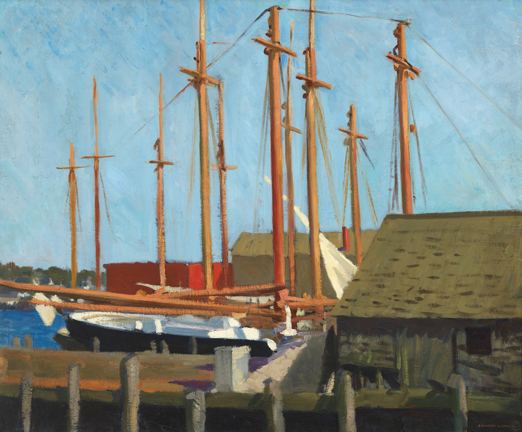 Several sailboats with tall wooden masts docked beside a weathered boathouse under blue sky.