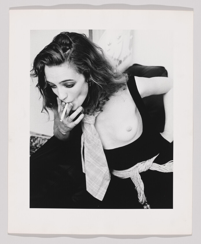 A woman smokes a cigarette while seated, wearing a loose tie and one breast exposed.